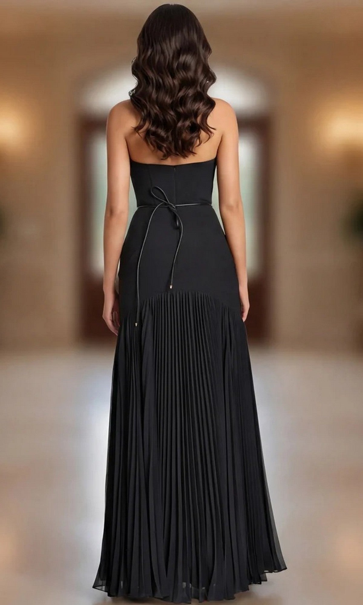 A woman with wavy brown hair is shown from behind, wearing the Jovani 47000 Long Formal Dress—a sleek black, strapless evening gown with a pleated skirt, a tie at the waist, and an elegant gold floral detail accentuating the waist, as she stands indoors.